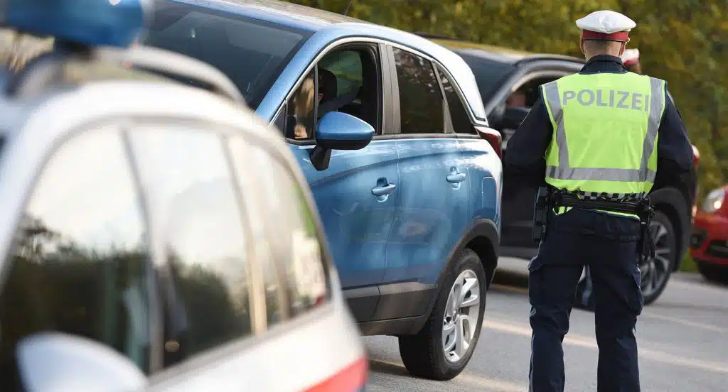 Foto von einer Grenzkontrolle, bei der ein Polizist von hinten zu sehen ist, der neben einem blauen Auto steht.